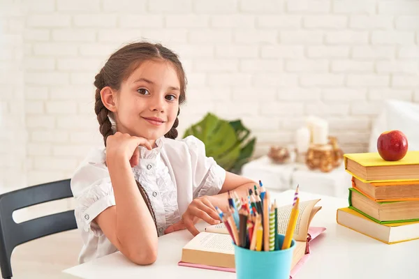 Радостная маленькая девочка, сидящая за столом с карандашами и учебником — стоковое фото