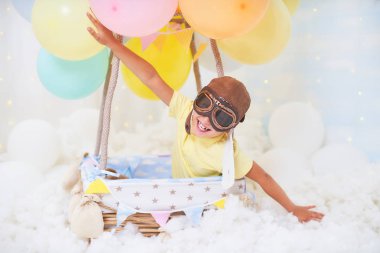 Küçük bir çocuk bulutların içinde balon sepeti içinde oturuyor. 