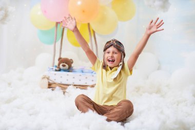 Küçük bir çocuk bulutların içinde balon sepeti içinde oturuyor. 