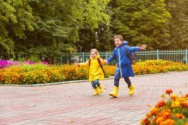 Niños felices ríen, se apresuran y corren a la escuela, vestidos con Raincoa —  Fotos de Stock