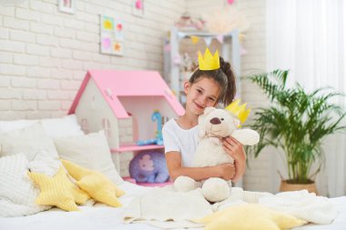 7 yaşında, kafasında kağıt bir taçla sabahın köründe yatakta oturan komik küçük bir kız. Çocuk mutlu bir şekilde gülümsüyor ve çocuk odasında oyuncak ayıya sarılıyor. Oyuncak ayı onun arkadaşı..