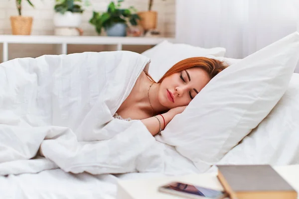 Attractive red-haired beauty sleeps with both hands under her head on a pillow, a book and a mobile phone are on table. cute girl sleeping in the bedroom in the morning light. healthy, sound sleep.