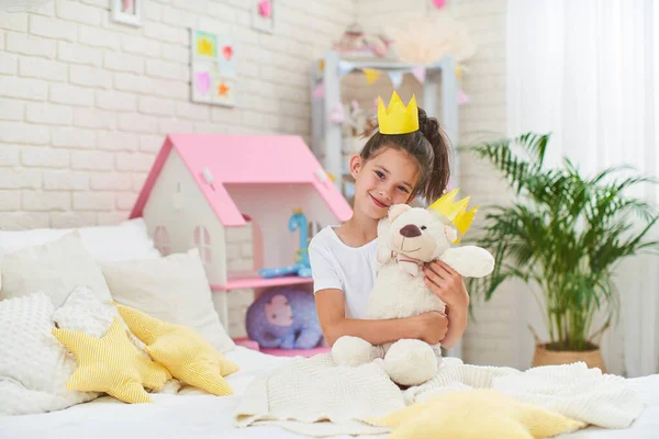有趣可爱的小女孩7岁 早上坐在床上 头上戴着纸冠 孩子们在孩子们的房间里开心地笑着 抱着一只玩具熊 泰迪熊是她的朋友 — 图库照片
