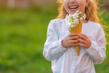 Gün batımında, gülen küçük bir kız tarafından bulanık doğal bir arka plan karşısında tutulan vişne çiçekleriyle dolu waffle külahının yakın çekimi. Baharı kutlama konsepti.