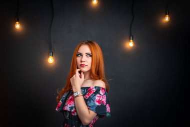 Çiçek desenli siyah elbiseli güzel, çarpıcı kızıl saçlı kadın, parlak ışıklarla siyah arka planda duruyor. Akşam makyajı ve açık elbisesi olan çekici bir genç kız başka yere bakıyor..