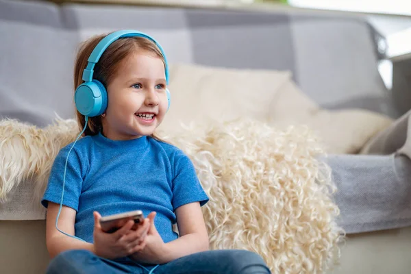 Happy little girl listening to music in blue headphones, at home. The child is sitting with smartphone in his hands and enjoys listening to his favorite songs and singing. Funny emotions. Copy space.