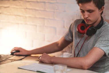 Boynuna kulaklık takmış, ödev yapıyor, seans yapıyor. Dizüstü bilgisayar kullanarak ve notlar alarak. konsept çevrimiçi öğrenme, sınavlara hazırlık. Sosyal uzaklık, kendini izole etme..