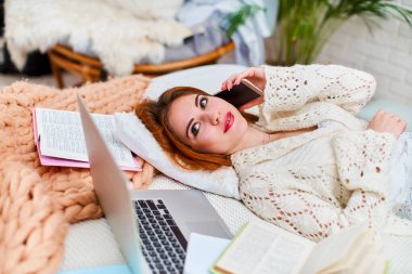 Sevimli genç kız ev ödevi yapmak yerine yatağında akıllı bir telefonla yatıyor. Bir öğrenci internetten koronavirüs karantinasında dizüstü bilgisayarla, uzaktan öğrenimle, kendi kendini eğitmeyle öğrenim görür..