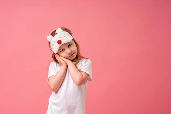 An adorable little girl in a sleep mask put her hands under her cheek on a pink background. The child wants to sleep. Bedtime. The concept of healthy sleep. Copy space.
