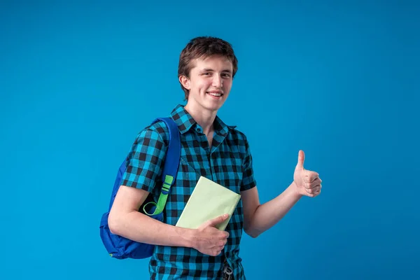 Smiling young man student in casual clothes with backpack holding books isolated on blue background. High school education is concept of University College. Layout of copy space. I give him thumbs-up.