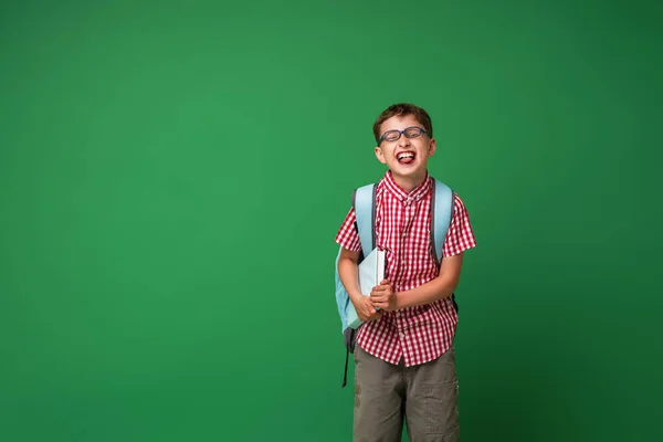 本とバックパックを持ってメガネを持ついたずら笑いの学童7歳 緑の背景に隔離された 学校に戻る — ストック写真
