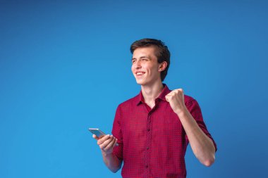 Mavi arka planda cep telefonu olan gülümseyen yakışıklı bir adam. Heyecanlı genç adam, ileriye bakıyor ve seviniyor, başarıyı kutluyor, akıllı telefonu elinde tutuyor. Zafer, indirim, kazanç.