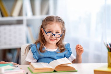 Okula dönün! Gözlüklü mutlu küçük bir kız odada bir kitapla bir masada oturuyor. Çocuk okul için hazırlanıyor. Ödevini yap. Evde eğitim kavramı.