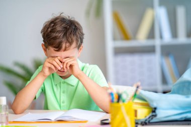 Yorgun okul çocuğu ağlıyor, gözlerini ovuşturuyor çünkü artık zorbalık yapmak ya da okumak istemiyor. Ev ödevi. Oğlan gözlerini yorgunlukla ovuyor, kitap ve ders kitapları okuyor. görme sorunu.