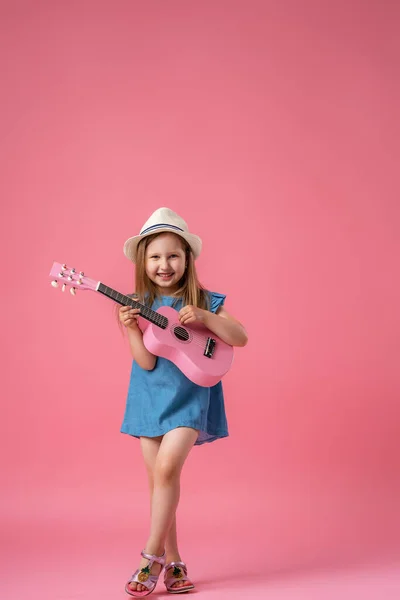 一个快乐的白人小女孩 穿着短短的斜纹棉布衣服 头戴一顶帽子 弹着粉红的Ukulele吉他 孩子在粉色的背景下弹奏乐器和唱歌 — 图库照片