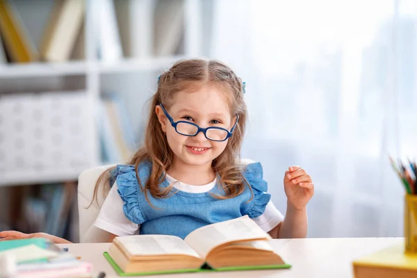 学校に戻って 部屋に本があるテーブルには眼鏡をかけた幸せな小さな女の子が座っている 子供は学校の準備をしている 宿題をしろ 家庭教育の概念 — ストック写真