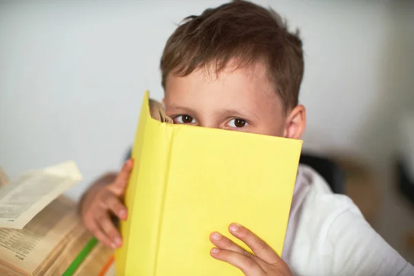 眼鏡をかけた賢い少年は黄色い本の後ろに顔を隠し宿題をしながらカメラを見る — ストック写真