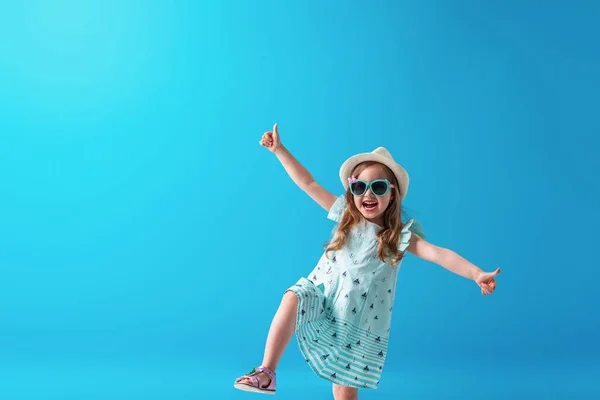 一个可爱的小女孩 身穿连衣裙 头戴帽子 戴着太阳镜 摆出一副蓝色背景的样子 孩子们会很高兴开始暑假 然后去旅行 — 图库照片