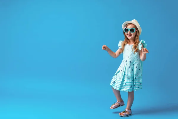 4歳の可愛い女の子が青い背景にドレスと帽子とサングラスをかけてポーズをとっています 子供たちは夏休みを開始し 旅行に行くことが幸せになります — ストック写真