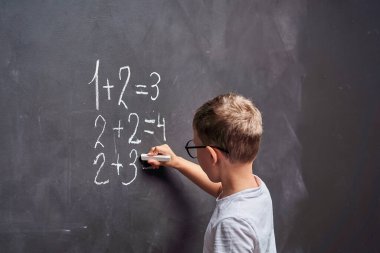 İlkokul eğitimi. Yan görüş öğrencisi matematik dersinde bir tahtada matematik örneği çözüyor. Bir ilkokul öğrencisi karatahtanın başında duruyor ve bir örneğe cevap yazıyor, cevabı düşünüyor..