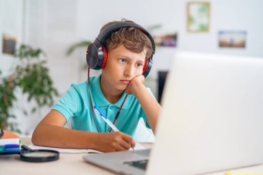 Kulaklıklı üzgün beyaz çocuk öğretmeniyle video görüşmesi yapmak için dizüstü bilgisayar kullanıyor. Çocuk uzak mesafe öğrenme biçiminden bıktı. E-Eğitim Mesafesi Öğrenme, Ev Eğitimi.