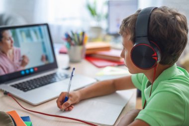 Küçük çocuk öğretmeniyle video görüşmesi yapmak için dizüstü bilgisayar kullanıyor. Ekranda, dersten konuyu açıklayan öğretmenle çevrimiçi bir ders gösteriliyor. E-Eğitim Uzaklık Öğrenimi, Ev Eğitimi. Yan görünüm.