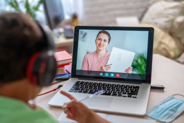 Küçük çocuk öğretmeniyle video görüşmesi yapmak için dizüstü bilgisayar kullanıyor. Ekranda, dersten konuyu açıklayan öğretmenle çevrimiçi bir ders gösteriliyor. E-Eğitim Uzaklık Öğrenimi, Ev Eğitimi. Omzunun üstünden.