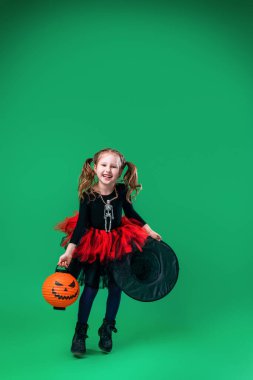 Mutlu Cadılar Bayramı! Elinde balkabağı ve şapkası olan sevimli küçük cadı yeşil arka planda zıplıyor. Çocuk Cadılar Bayramı için hazırlanıyor. Çocuk elinde balkabağı şeklindeki fenerle korkmuş. Hile ya da izleme