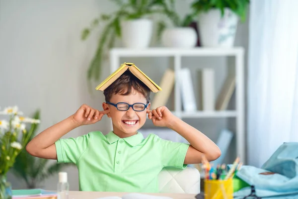 眼鏡をかけた面白い少年と頭の上の本が耳を閉じてテーブルに座っていました レッスンの準備学校に戻ります 子供は疲れて勉強したり遊んだりだましたり — ストック写真