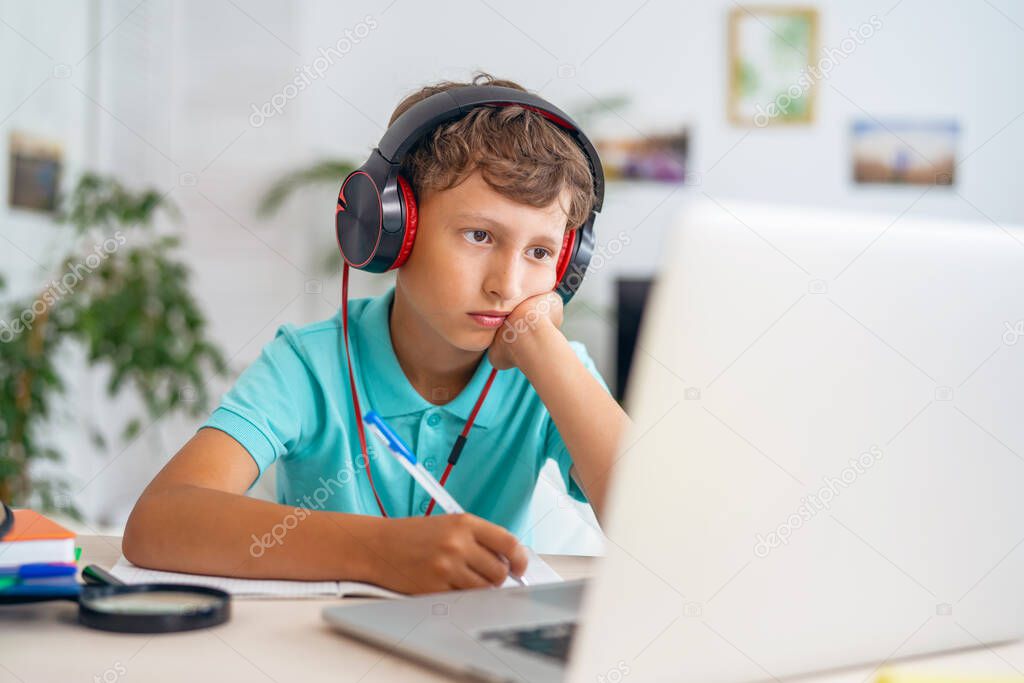 sad Caucasian boy in headphones uses a laptop to make a video call with his teacher. The child is tired of the form of distance learning. E-Education Distance Learning, Home Schooling.