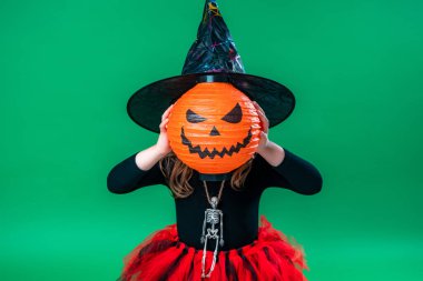 Mutlu Cadılar Bayramı! Yeşil arka planda balkabağı olan şirin bir cadı. Çocuk Cadılar Bayramı için hazırlanıyor. Çocuk elinde balkabağı şeklindeki bir fenerle korkuyor. Hile ya da treak