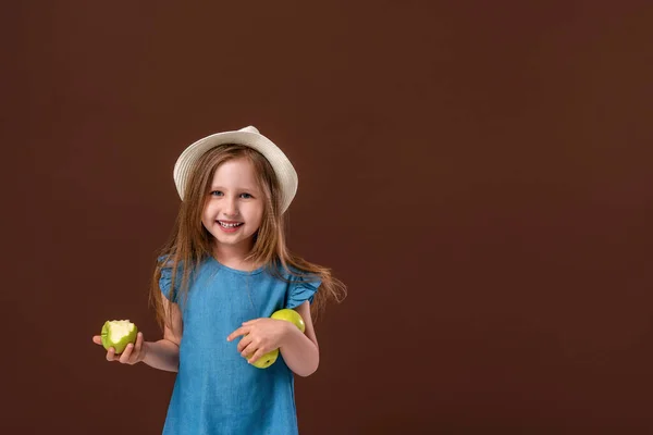 一个可爱的小女孩抱着一个棕色背景的苹果的肖像 那孩子戴着草帽 吃着苹果 收集秋天的收获 吃水果 秋天的收获 — 图库照片