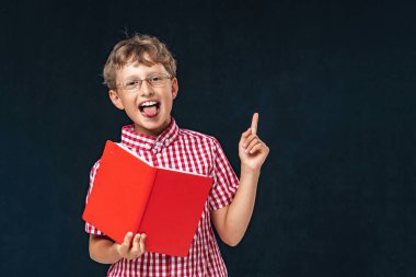Okula dönelim. Gözlüklü, dili elinde kitapla gülümseyen, siyah bir arka planda poz veren komik bir çocuk. Okul için hazırlanıyor, tahtada arka planda. Eğitim.