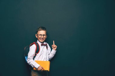 Neşeli, yaramaz, beyaz çocuk elinde bir kitap tutuyor, tahtada duruyor. Siyah arka planda heyecanlı bir çocuk dikkatini boş alana yöneltir. Okula geri dön..