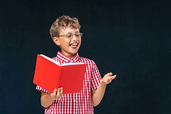 学校に戻る グラスと本を手にした面白い笑顔の少年が黒い背景にポーズをとっています 黒板を背景に学校への準備 子供は数え方を学び — ストック写真