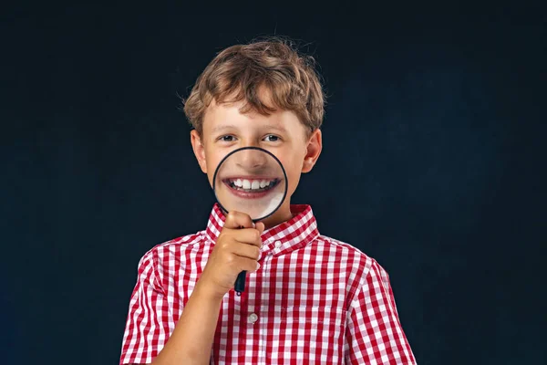 黒を背景に口の近くに虫眼鏡をかけた面白い子 少年は虫眼鏡を通して歯を見せる 歯の矯正 小児用整形外科医 — ストック写真