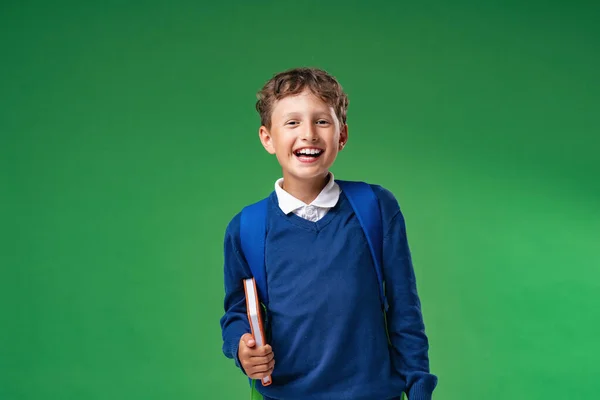 7年間制服姿のいたずら笑い少年が本やリュックを持っています 緑の背景に隔離された 学校に戻る 子供はとても興奮して喜んでいます 訓練を始める — ストック写真