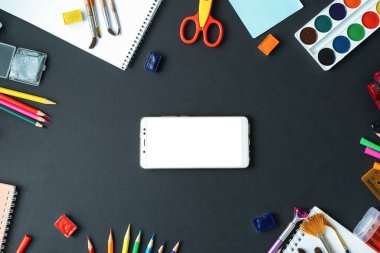 Okul malzemeleri kurulun siyah arka planında. Merkezde, siyah bir yüzey üzerinde beyaz ekranlı bir akıllı telefon var. Suluboya resimler, kalemler, keçeli kalemler, fırçalar ve çizim malzemeleri..