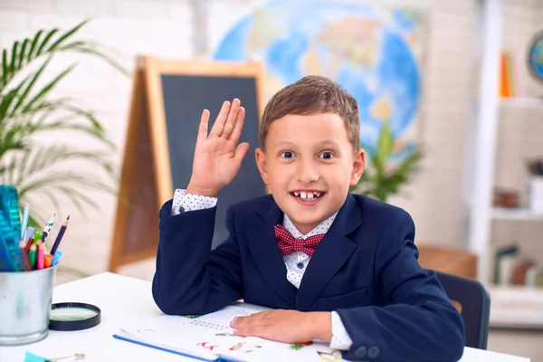 好奇的小男孩穿着学校的课本 一个穿制服的孩子坐在桌旁举手回答 以学校课堂为背景 准备上学了回学校去 — 图库照片