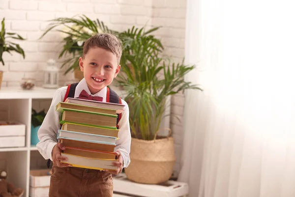 かわいい探究心のある少年は学校の教科書を着ている 彼の手に本の山と一様に子供 背景にホームインテリア 学校の準備だ 学校に戻る — ストック写真