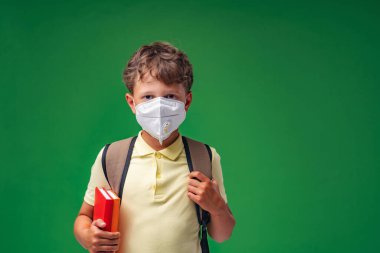 Sırt çantalı, koruyucu maskeli, yeşil arka planda duran üzgün okul çocuğu. Çocuk okula üzgün gidiyor, maske takmak istemiyor. Grip ve koronavirüs salgınları sırasında korunma