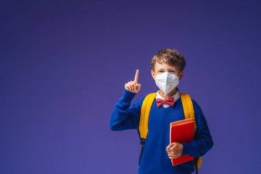Koruyucu maskeli okul çocuğu, üniformalı, mor arka planda kitap ve sırt çantalı. Heyecanlı çocuk parmağını kaldırır ve dikkatleri boş alana yönlendirir. Okula dönelim. salgın koronavirüsler