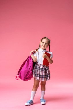 Sırt çantası ve elinde not defteriyle mutlu, gülümseyen küçük bir kız stüdyoda pembe bir arkaplanda poz veriyor. Bu çocuk ilk kez okula gidiyor. Okula dönelim. Boşluğu kopyala