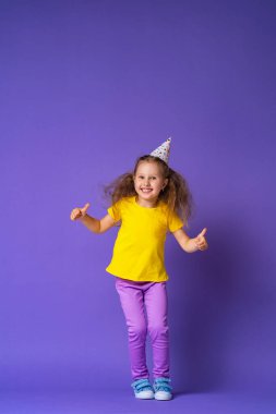 Mutlu yüzlü bir tatil şapkası takan küçük tatlı kız mutlu, atlıyor ve mor arka planda bir onay, bir sınıf işareti gösteriyor. Çocuğun doğum günü. Boşluğu kopyala