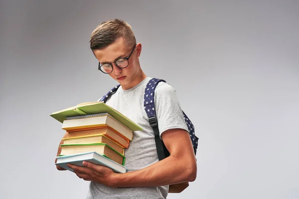 Portrait of a smart student in glasses with a backpack and a stack of books in his hands on a gray background.The guy is reading a book enthusiastically. Gain knowledge. Getting higher education.