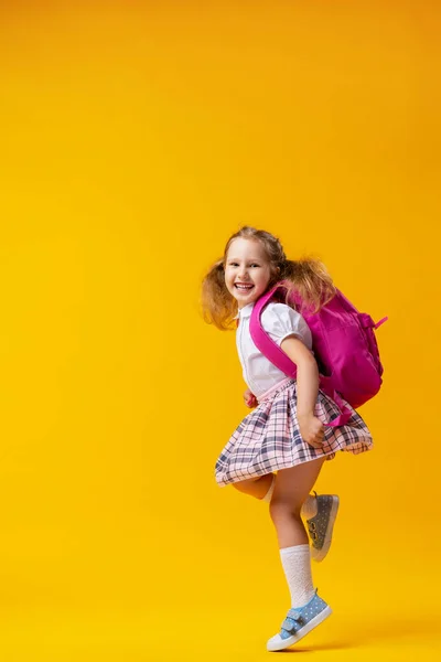 穿着校服的活泼淘气的女学生背着背包跳到黄色的背景上 生动的画面又回到了学校的概念 假期开始了 回学校去男孩准备好要学习了 — 图库照片