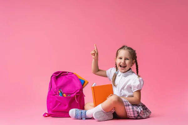 穿着制服 背着背包和书本 快乐地微笑着的小女孩坐在演播室的粉红背景上 那孩子把手指尖了一下 回学校去复制空间 — 图库照片