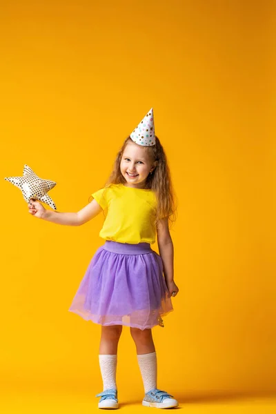 生日快乐 一个戴着节日礼帽 背景为黄色的小女孩的工作室肖像 那孩子笑了 手里拿着一个纺织明星 等待礼物 — 图库照片