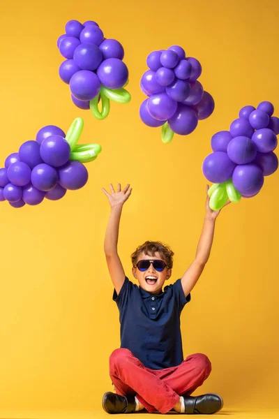 时尚男孩7岁 戴着时髦太阳镜 在黄色橙色背景下 用气球做成葡萄 孩子们在工作室摆姿势 用气球做成水果 夏天的概念 维生素 — 图库照片