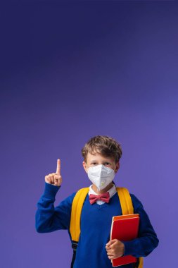 Koruyucu maskeli okul çocuğu, üniformalı, mor arka planda kitap ve sırt çantalı. Heyecanlı çocuk parmağını kaldırır ve dikkatleri boş alana yönlendirir. Okula dönelim. salgın koronavirüsler